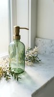 Green glass soap dispenser with brass pump on marble bathroom counter surrounded by eucalyptus branches in natural window light for spa-inspired home decor.