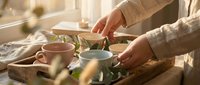 Woman in beige sweater preparing herbal tea on wooden tray with ceramic cups and teapot in warm sunlit kitchen for relaxation and wellness routine.