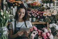 Young Asian female florist using digital tablet while working in flower shop, surrounded by fresh spring blooms including tulips, roses and daffodils in retail setting.