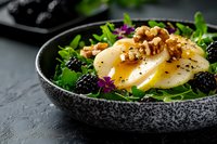 Fresh pear and blackberry salad with arugula, walnuts and edible flowers served in textured black ceramic bowl on dark stone background.