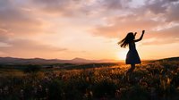 Woman with long dark hair in silhouette, wearing a dark dress, standing in a wildflower field during golden hour sunset, arms raised in celebration, mountains visible in distant background.