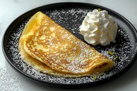 Thin golden crepe served on black plate with powdered sugar dusting and fresh whipped cream dollop, close-up view showing delicate texture and light browning.