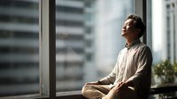 Asian man in meditation pose by window with urban cityscape view, peaceful mindfulness practice in modern apartment setting.