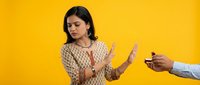 Young Indian woman in traditional patterned dress making stop gesture with hands, refusing cigarette offer on bright yellow background for health awareness campaigns.