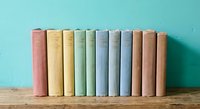 Row of vintage pastel colored books arranged on wooden shelf against turquoise wall. Literary collection for education, reading, library decor concepts.