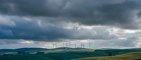 Wind turbines generating renewable energy on rolling green hills under dramatic stormy sky with dark clouds and sunlight breaking through atmosphere.