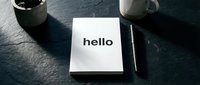 White notebook with bold hello text on dark slate desk beside pen, mug and plant, minimal workspace concept for branding and communication.