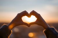 Hands forming heart shape against sunset sky with warm golden light shining through fingers creating romantic silhouette for love and affection concepts.