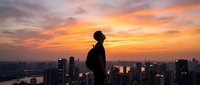 Young African American businessman silhouette standing on rooftop overlooking city skyline at golden hour sunset with dramatic orange clouds and urban landscape.