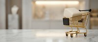Miniature shopping cart filled with cardboard box on white marble surface with blurred retail store background, representing e-commerce, online shopping, and delivery services.