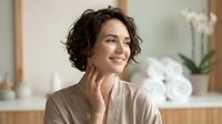 Woman with short dark wavy hair, fair complexion, wearing beige linen shirt in modern bathroom with natural light, touching face thoughtfully, skincare and wellness concept.