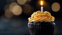 Chocolate cupcake with swirled orange buttercream frosting and gold sprinkles topped with lit birthday candle against dark background with festive bokeh lights.