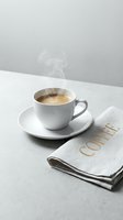 Steaming hot espresso in a white ceramic cup on a saucer beside a linen napkin with gold coffee lettering on a gray surface.