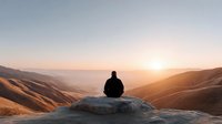 Person sitting on rock overlooking mountain valley at golden hour sunset, meditation and mindfulness concept for wellness and mental health content.