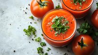 Fresh tomato gazpacho soup garnished with herbs and black pepper in glass cups, surrounded by ripe tomatoes on light textured background.