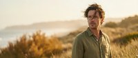Thoughtful middle-aged Caucasian man in olive shirt standing in golden hour coastal landscape with warm sunlight and natural vegetation backdrop.