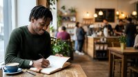 Young man with dark braided hair, African descent, wearing olive green sweater, writing in notebook at wooden table in busy cafe with warm lighting and blurred customers in background.