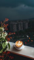 Latte art coffee cup with floral design next to red berries and white flowers on windowsill overlooking city lights at dusk, creating cozy atmosphere.