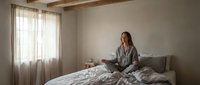 Young woman in gray pajamas sitting on unmade bed in cozy bedroom with natural light streaming through curtains, perfect for lifestyle and wellness content.