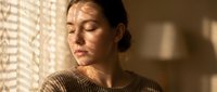 Young woman with closed eyes in peaceful meditation pose wearing cozy sweater in warm sunlit room with soft window light creating calming atmosphere.