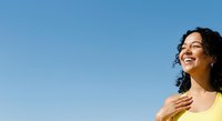Young woman with dark curly hair and medium skin tone wearing yellow tank top, smiling outdoors against clear blue sky. Portrait for lifestyle and wellness content.