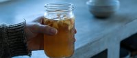 Person in gray sweater holding glass jar with iced tea or cold brew beverage outdoors on blue surface.