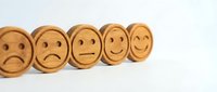 Five wooden coins with carved emoticons showing progression from sad to happy faces on white background for customer satisfaction surveys.