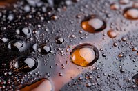 Water droplets on dark reflective surface with amber light shining through, creating dramatic macro photography effect with surface tension and reflection.