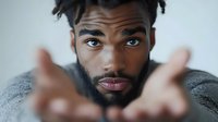 Young African American man with beard and modern hairstyle reaching towards camera with both hands, expressing emotional connection against light background.