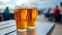 Two golden beer glasses with foam on outdoor table at sunset with blurred people in background for restaurant and pub advertising concepts.