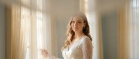 Young Caucasian woman in white lace dress standing by bright window with soft natural lighting creating warm atmospheric mood for lifestyle photography.