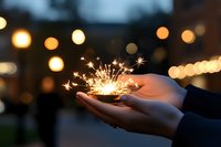 Glowing sparkler burning brightly in cupped hands against dark evening background with soft bokeh lights creating magical festive atmosphere.