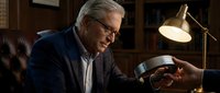 Mature man with gray hair and glasses, wearing dark blazer and light shirt, sitting at desk in home office with desk lamp and leather chair, focused on work or reading.