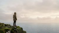 Solitary traveler in olive jacket standing on rocky cliff overlooking misty ocean horizon, contemplative outdoor adventure scene for travel and lifestyle content.