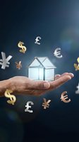 Hand holding transparent glass house model with floating currency symbols representing real estate investment and global property market opportunities.