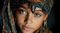 Young African woman with colorful braided hair and traditional headwrap looking directly at camera with striking eyes and natural makeup against dark background.