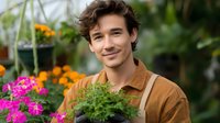 Young male gardener in brown jacket holding potted plants at greenhouse nursery, surrounded by vibrant pink and orange flowers, expressing genuine joy and satisfaction.