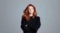 Young woman with red wavy hair wearing black sweater standing with crossed arms against gray background, expressing confidence and professional attitude.
