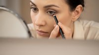 Young woman applying black eyeliner pencil while looking in mirror during morning beauty routine makeup application process in bathroom.