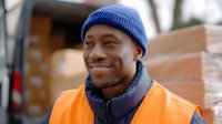 African American male warehouse worker wearing orange safety vest and blue beanie smiling confidently outdoors near delivery truck and cardboard boxes. Professional logistics portrait.