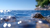 Crystal clear Mediterranean waters with smooth pebbles and rocks creating peaceful coastal scene. Shallow depth of field with bokeh light reflections on calm sea surface.