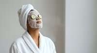 Young woman relaxing with facial mask and cucumber slices on eyes wearing white bathrobe and towel for spa skincare routine and wellness treatment.
