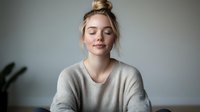 Young blonde woman with closed eyes in cozy gray sweater practicing mindfulness meditation at home. Peaceful relaxation and mental wellness concept.