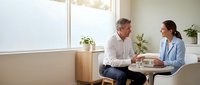 Middle-aged man and woman in business casual attire having a professional conversation in a bright office setting with plants and natural light from windows.