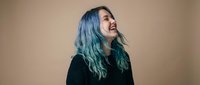 Young woman with vibrant blue green hair laughing joyfully against neutral beige background. Authentic happiness and positive emotions concept.