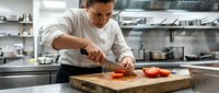Chef in white chef's coat and glasses slicing fresh tomatoes on wooden cutting board in professional kitchen for culinary, cooking, and food preparation content.