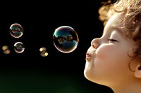 Young child with curly hair blowing soap bubbles against dark background, capturing magical moment of childhood play and wonder in golden sunlight.