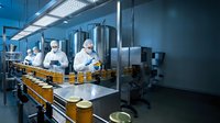 Food production workers in protective suits operating automated bottling line in modern sterile manufacturing facility with stainless steel equipment.