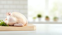 Raw whole chicken on wooden cutting board with fresh herbs in bright kitchen setting, selective focus on poultry against blurred background.