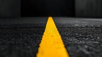 Bright yellow line painted on dark asphalt road surface with selective color effect, close-up view showing textured detail and perspective leading into distance.